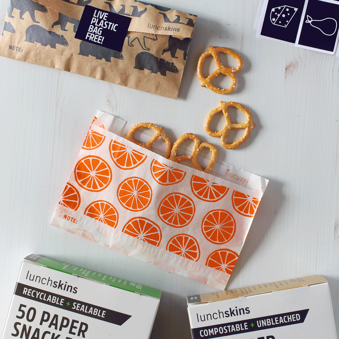 A styled flat-lay showing lunchskins branded paper snack bags and boxes on a white surface, with pretzels placed around them. The central bag has an orange slice print, while the top bag has dark animal silhouettes and the bottom boxes are white with green and black text.