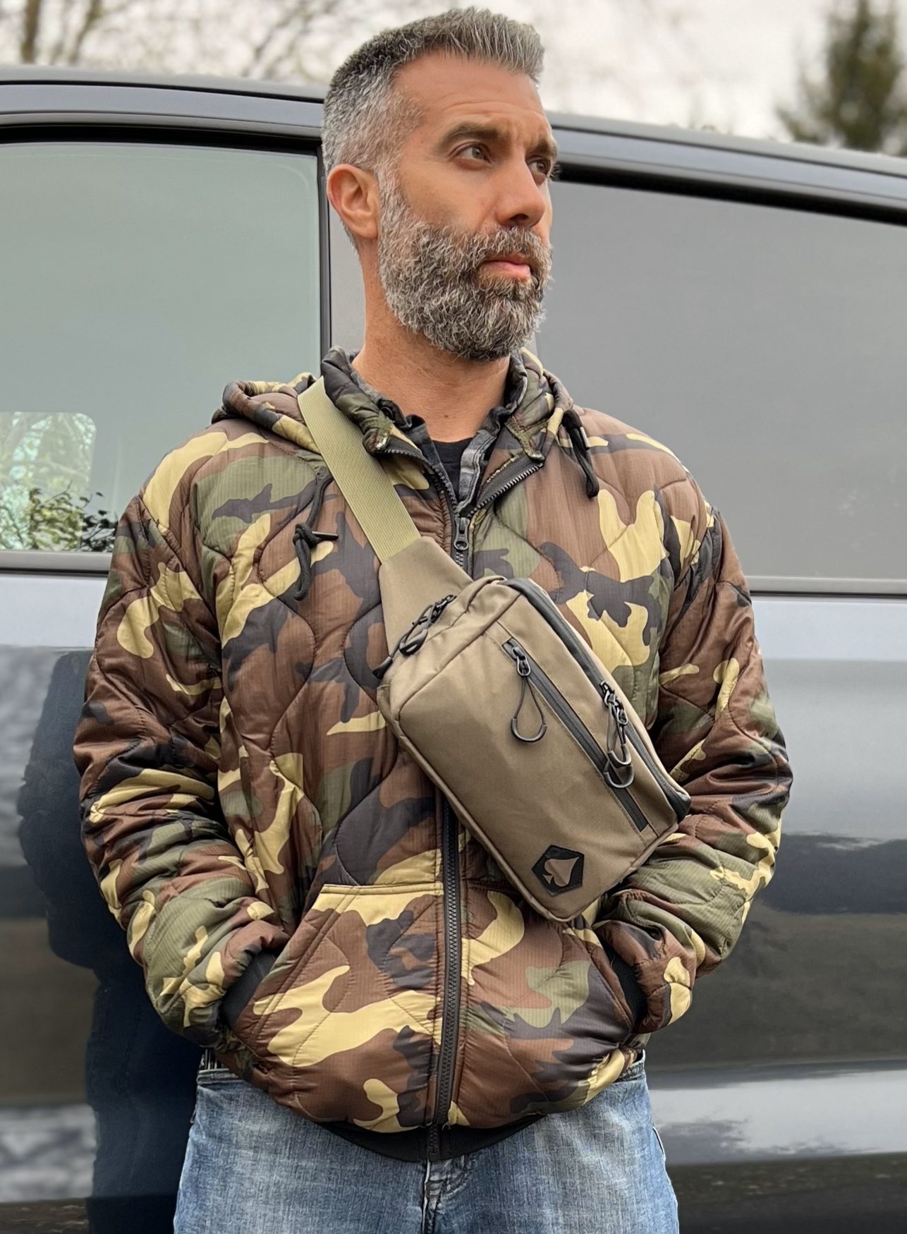 Man wearing a camouflage jacket with a crossbody bag standing next to a vehicle.
