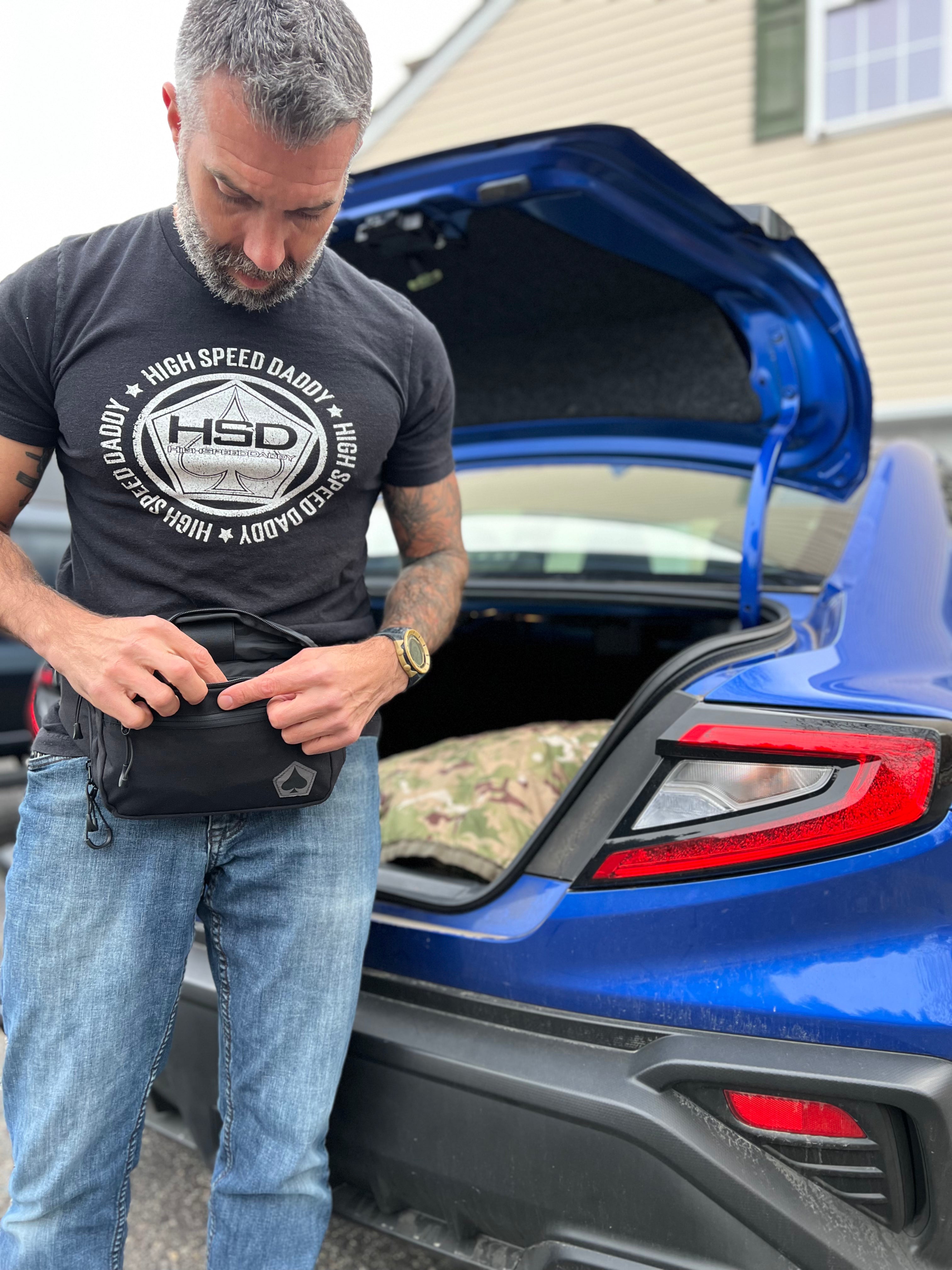 Man opening a blue car trunk with a black bag, wearing a gray t-shirt with 'High Speed Daddy' text.