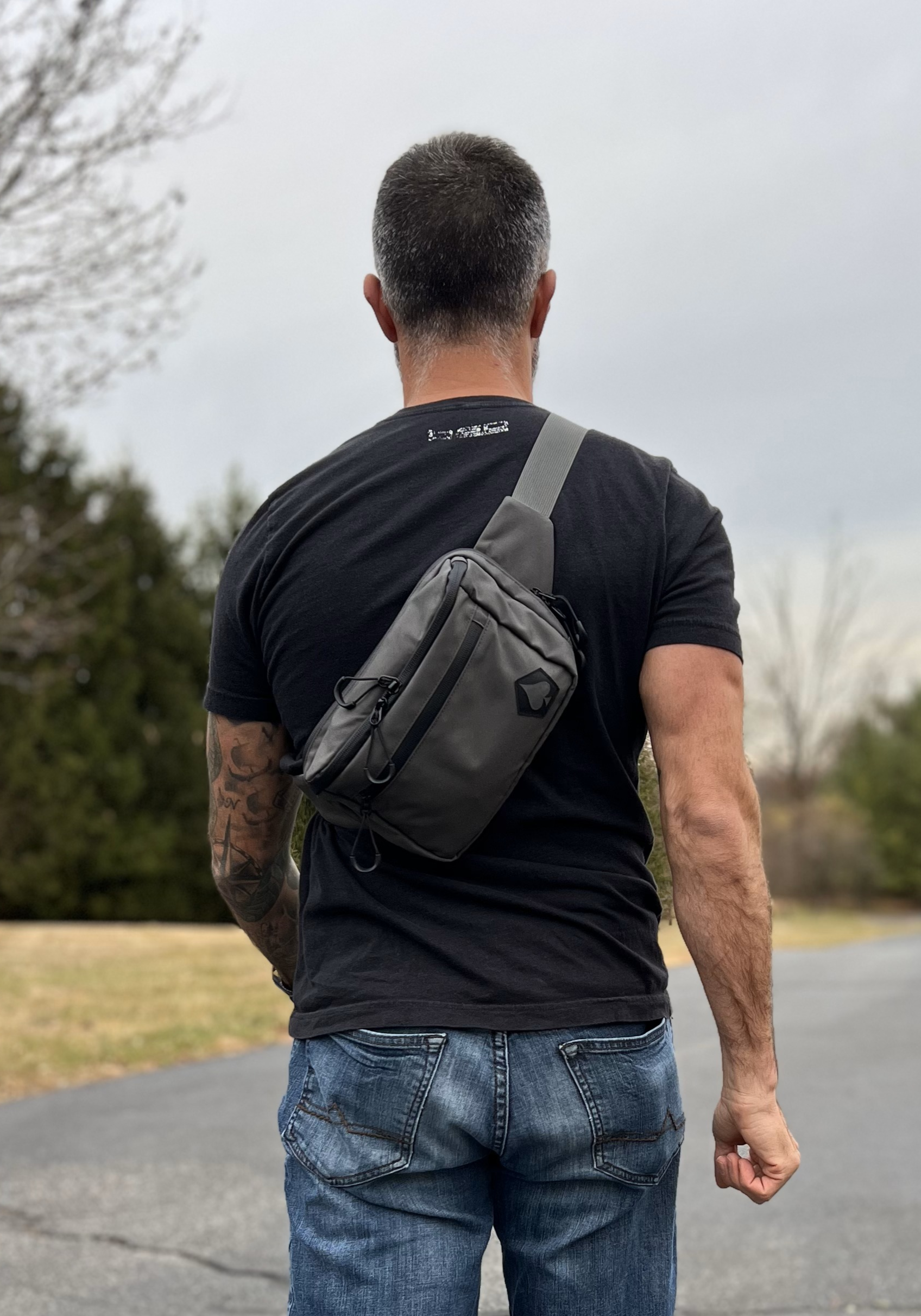 Man wearing a black t-shirt and jeans with a gray crossbody bag outdoors.