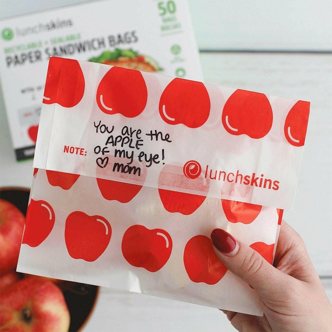 Lunchskins paper sandwich bag with apple design and 'You are the apple of my eye! Mom' text, held by a hand.