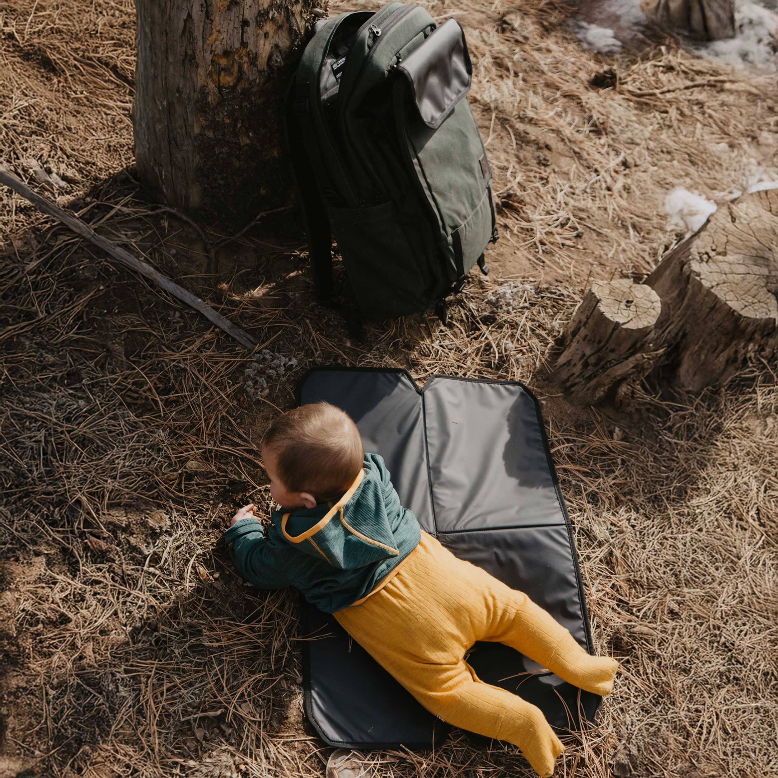 Child lying on a black mat in a forest setting with a green backpack nearby.