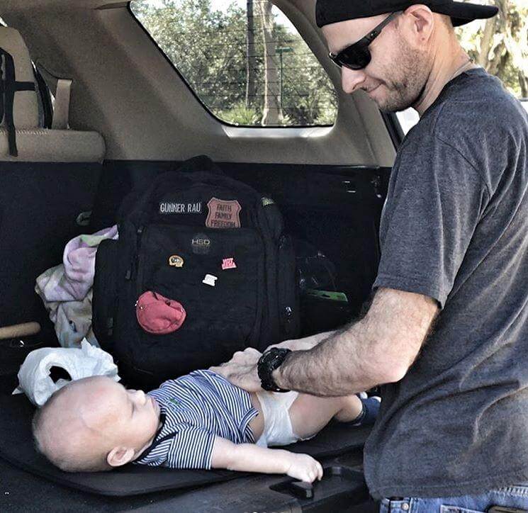 Man adjusting a baby in a car seat with a black bag and baby items in the back of a vehicle.